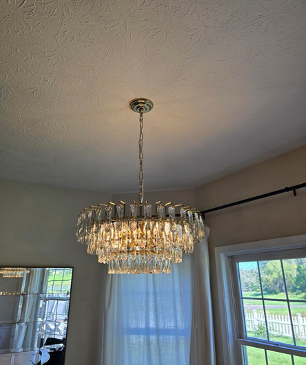 Crystal chandelier hanging from a ceiling in a bright room with windows and white curtains.