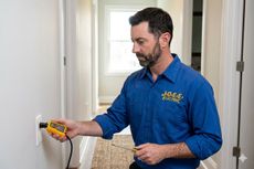 Technician in blue shirt tests an electrical outlet with a voltage tester in a hallway.