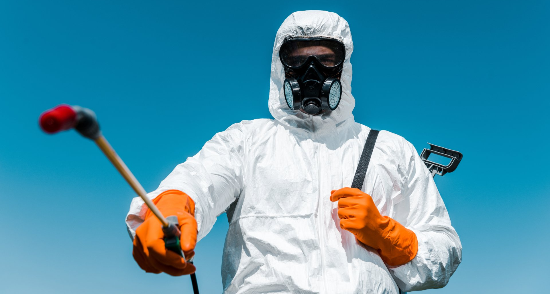 Technicien en combinaison blanche avec masque respiratoire et gants orange tenant un pulvérisateur.