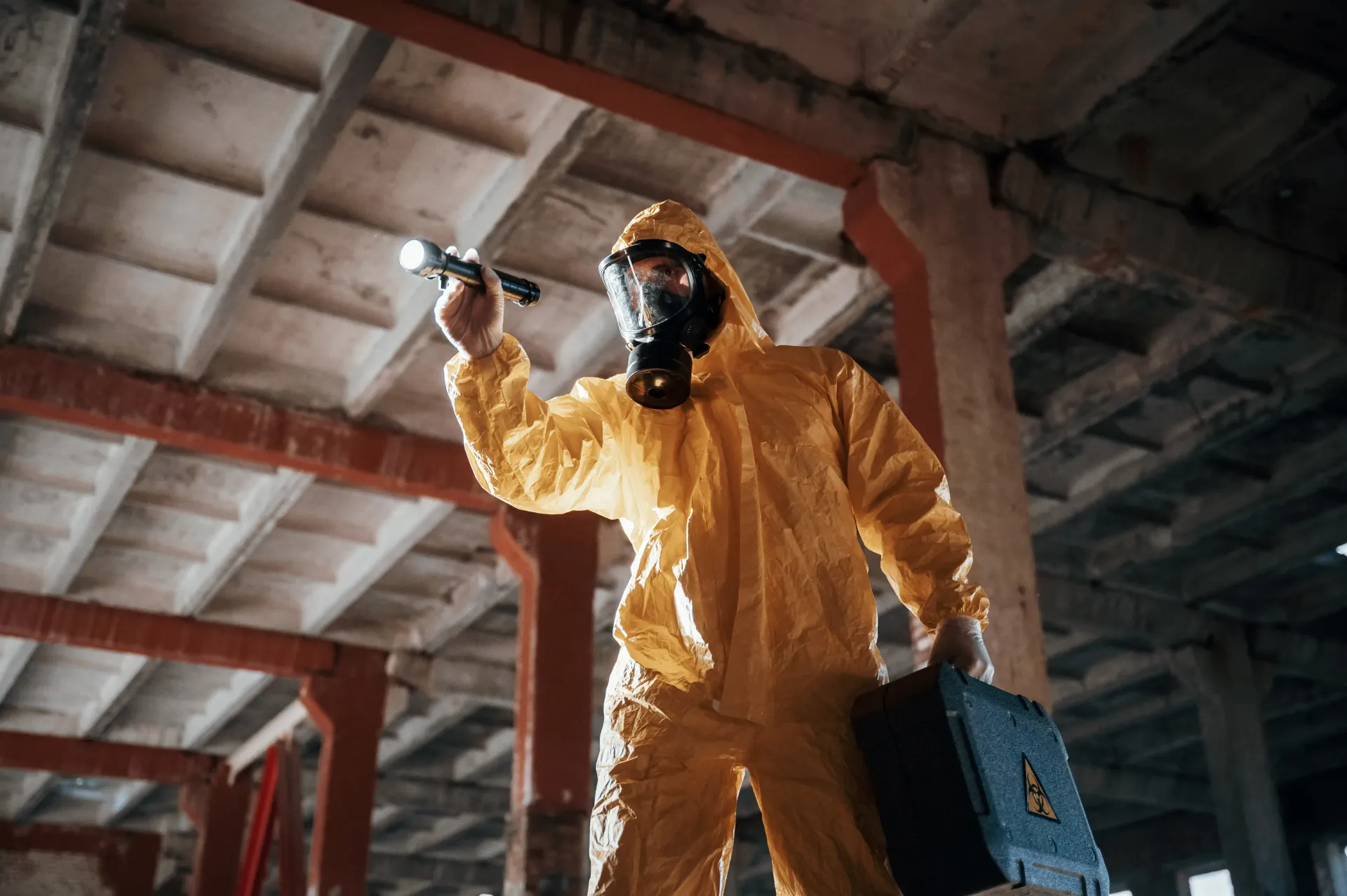 A man in a yellow protective suit and gas mask is holding a flashlight and a toolbox.