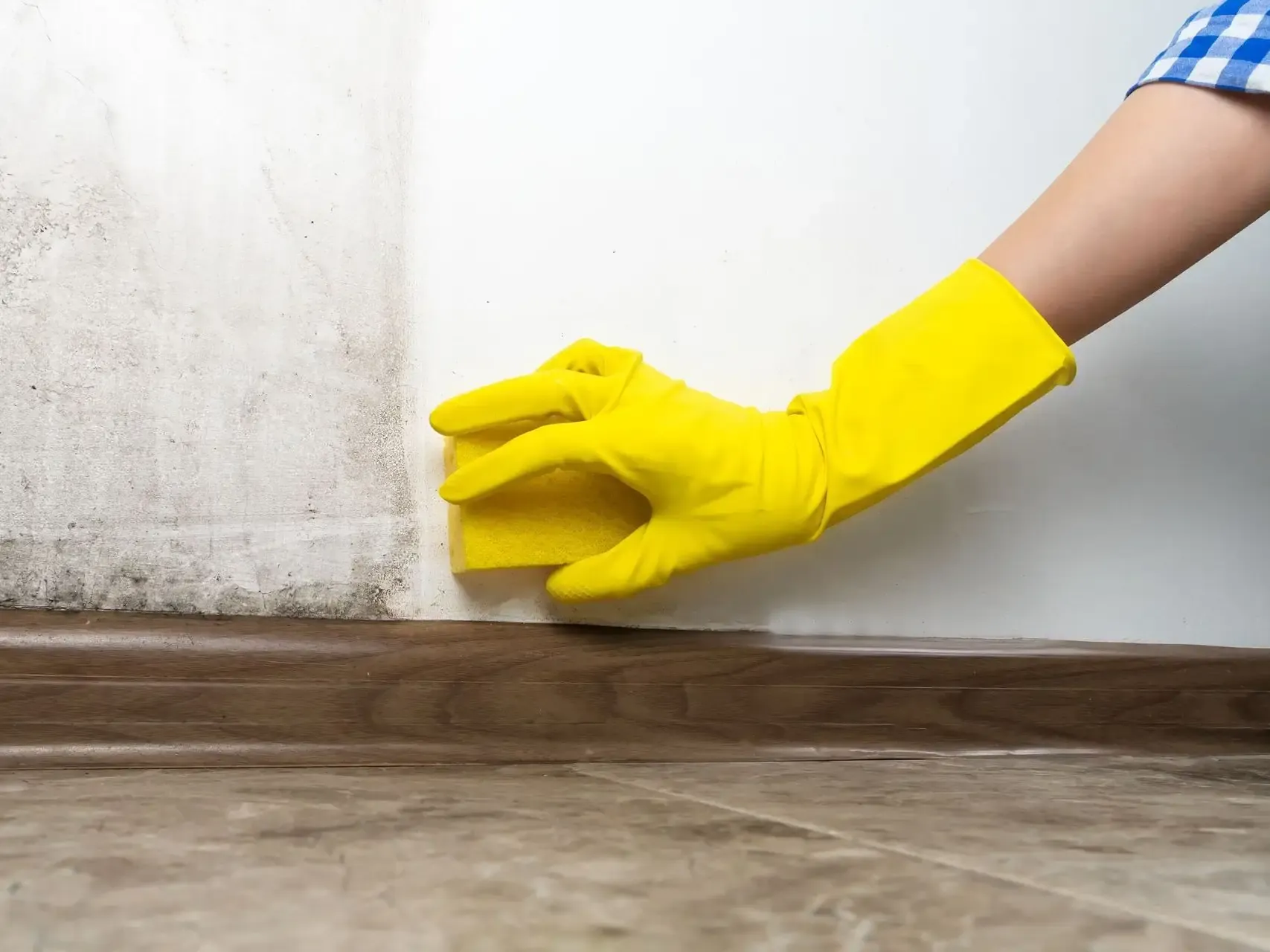 Yellow-gloved hand cleaning the wall-floor corner with a cloth, removing dirt from a stained surface.