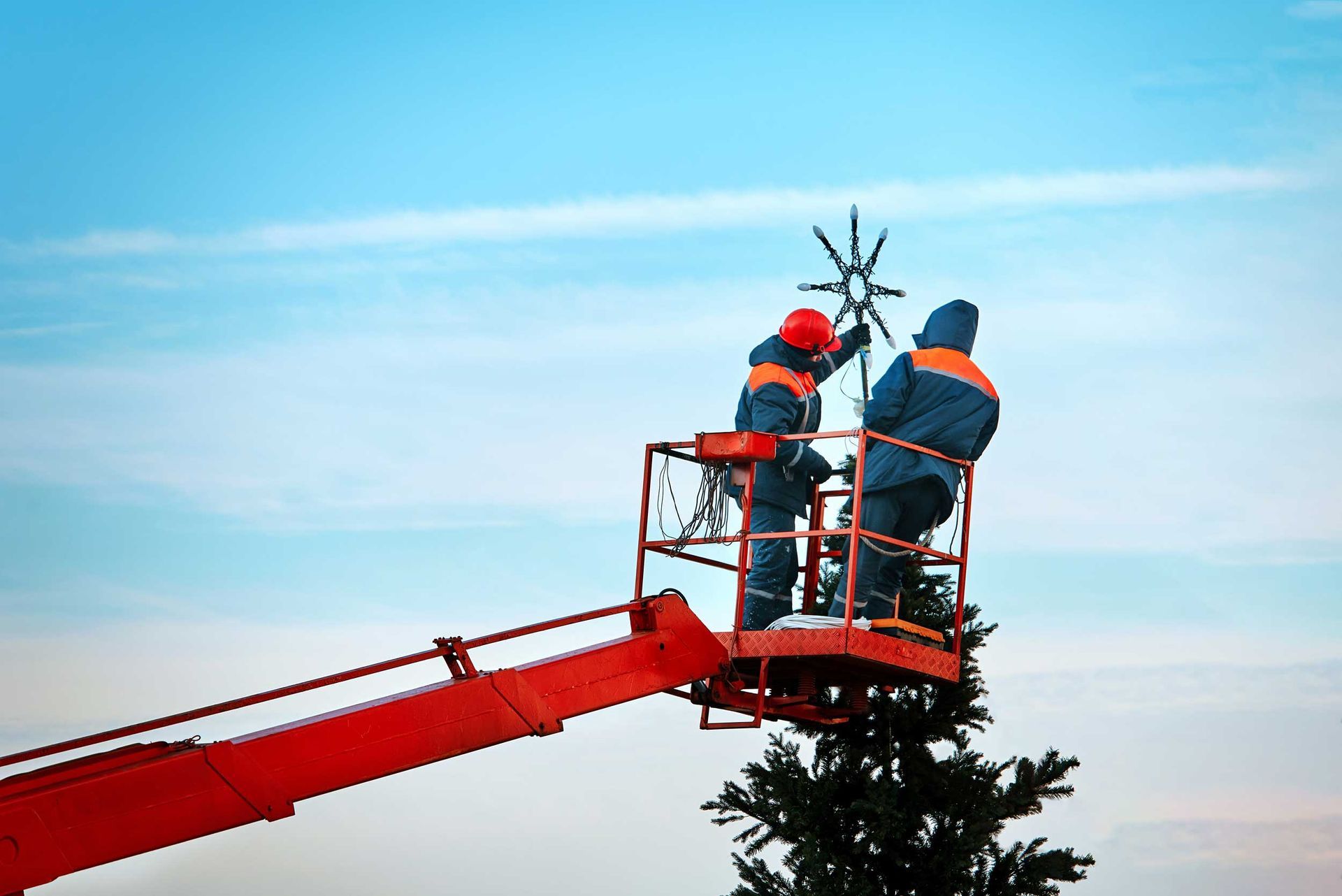 Two workers in orange safety gear on a red cherry picker trimming a tall tree against a blue sky
