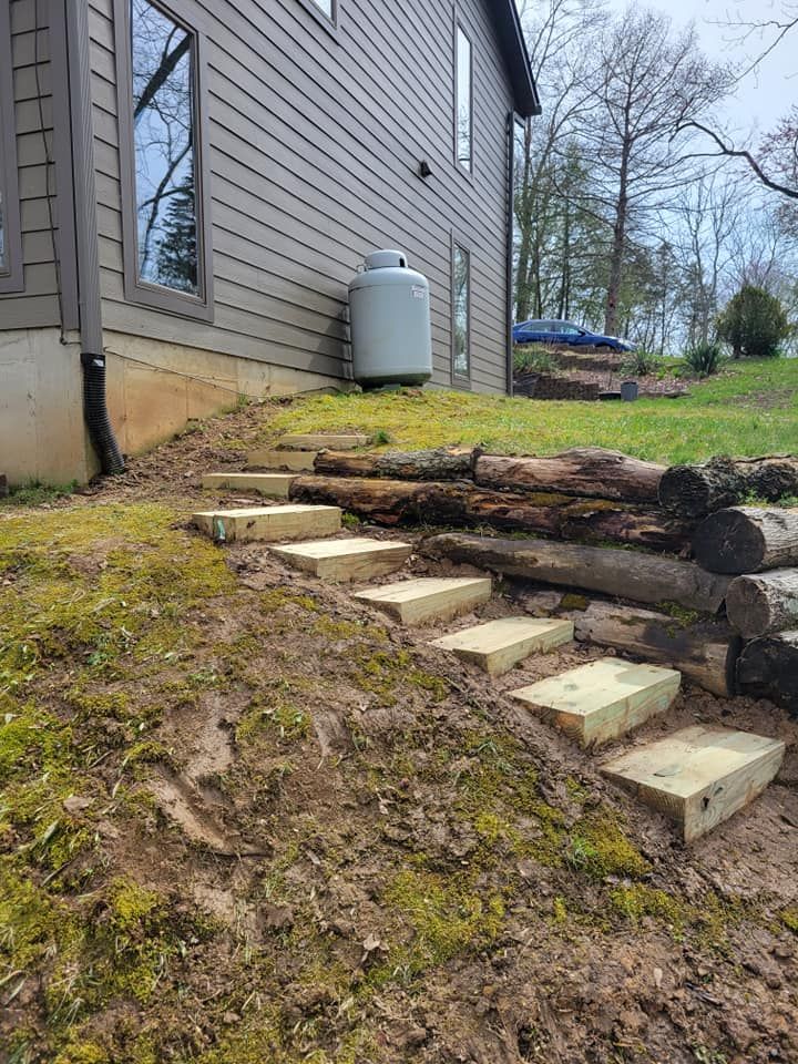 Side yard with sloped grass, stone steps, retaining logs, and a gray house wall with a gas tank.