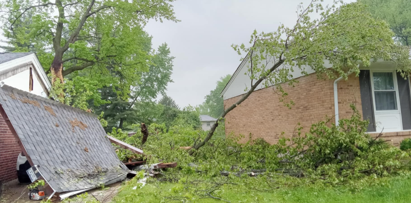 Storm damage in a yard, with a fallen tree, broken fence, and a damaged house roof.