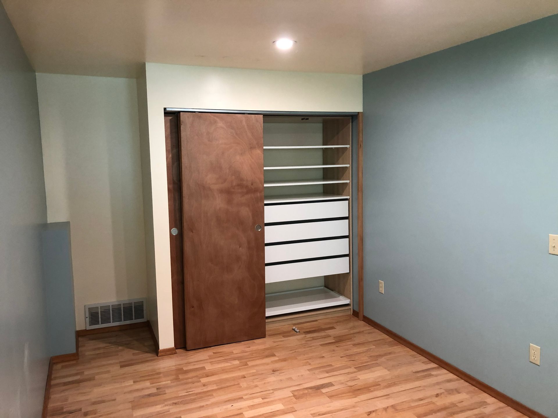 Empty room with light blue walls, wood floor, and an open closet with shelves.