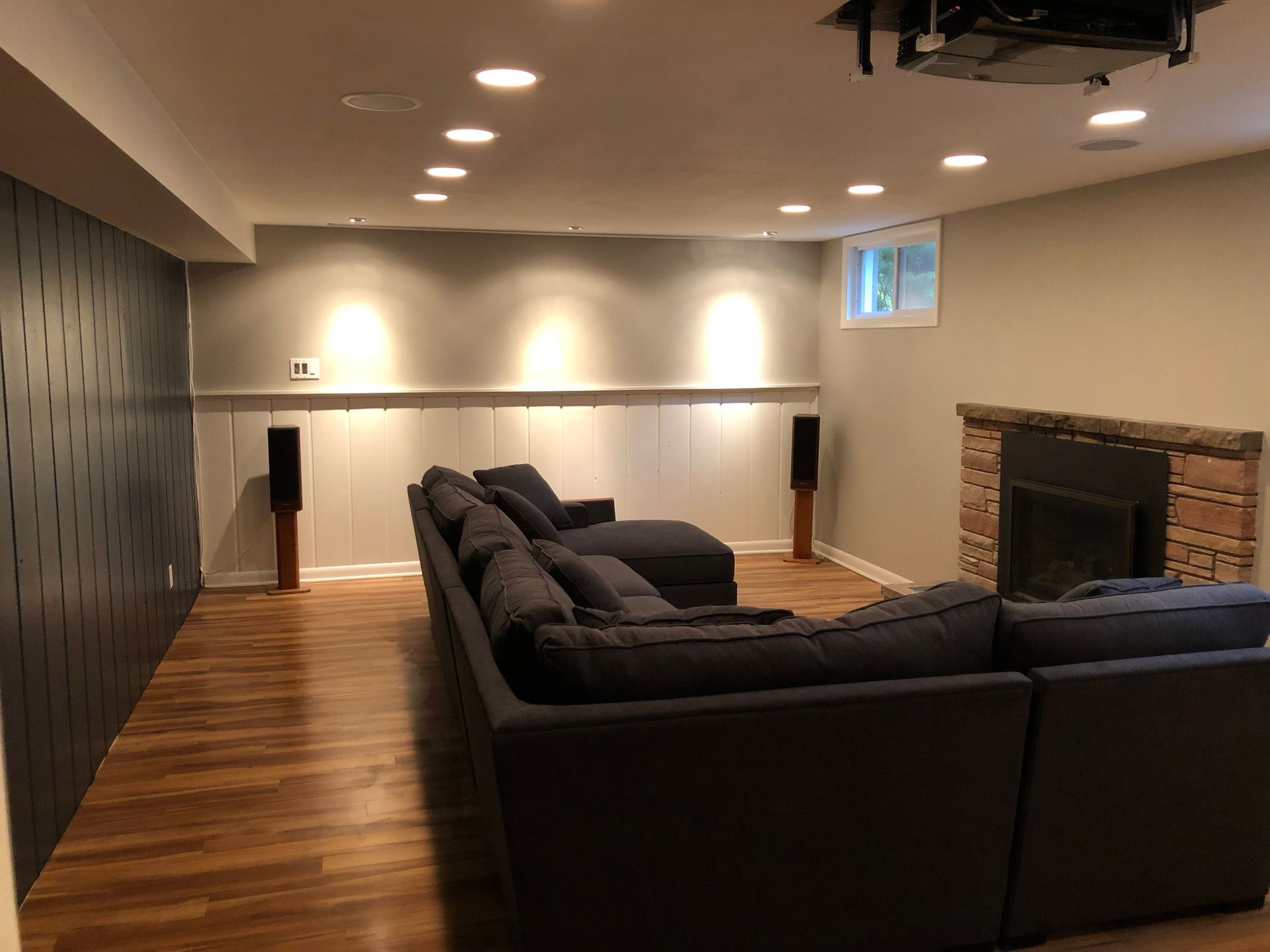 Spacious basement lounge with dark sectional sofa, wood floor, recessed lighting, and brick fireplace.