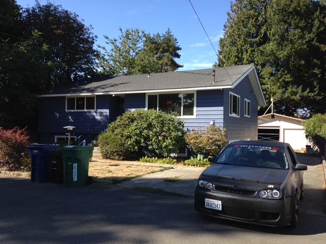 Blue house with parked black car and green trash bins in a sunny driveway.
