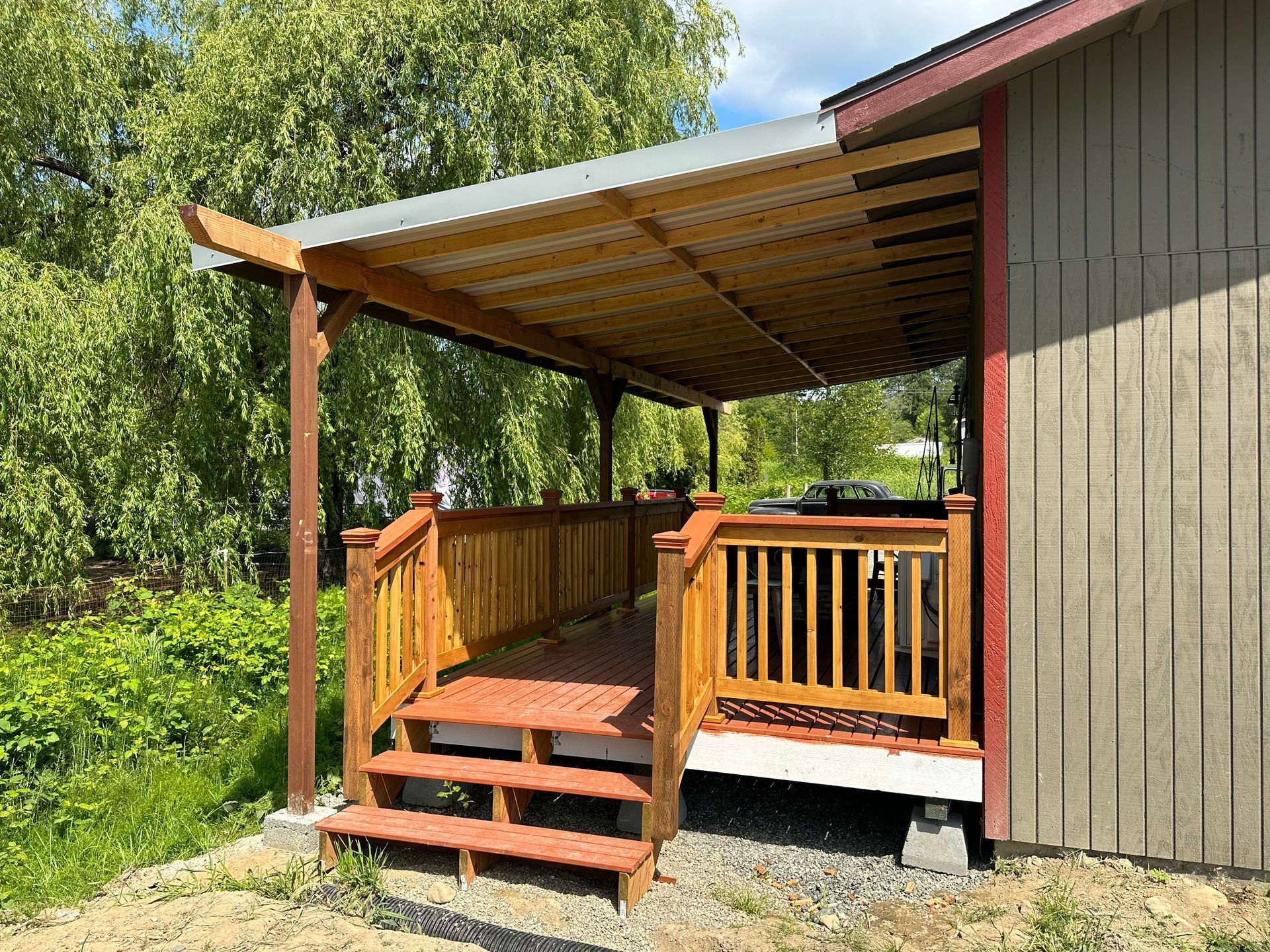 Wooden porch with steps and railing attached to a gray house, covered by a slanted roof