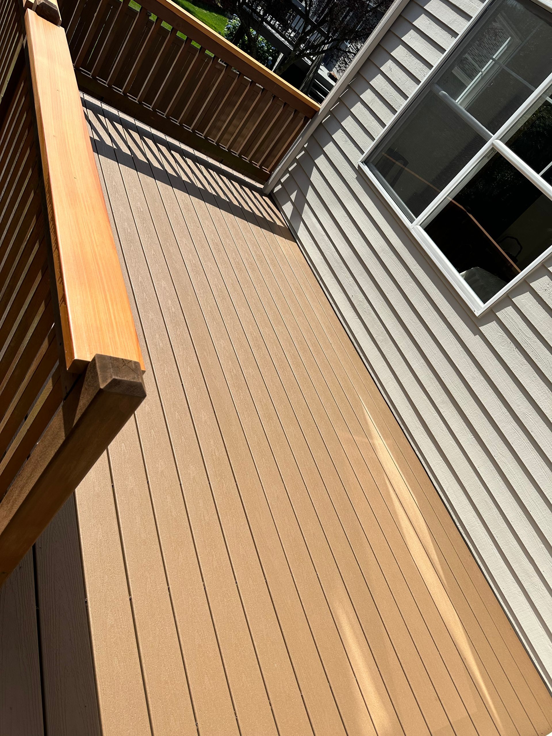 Sunlit wooden deck beside a house with railing and window, viewed at an angle