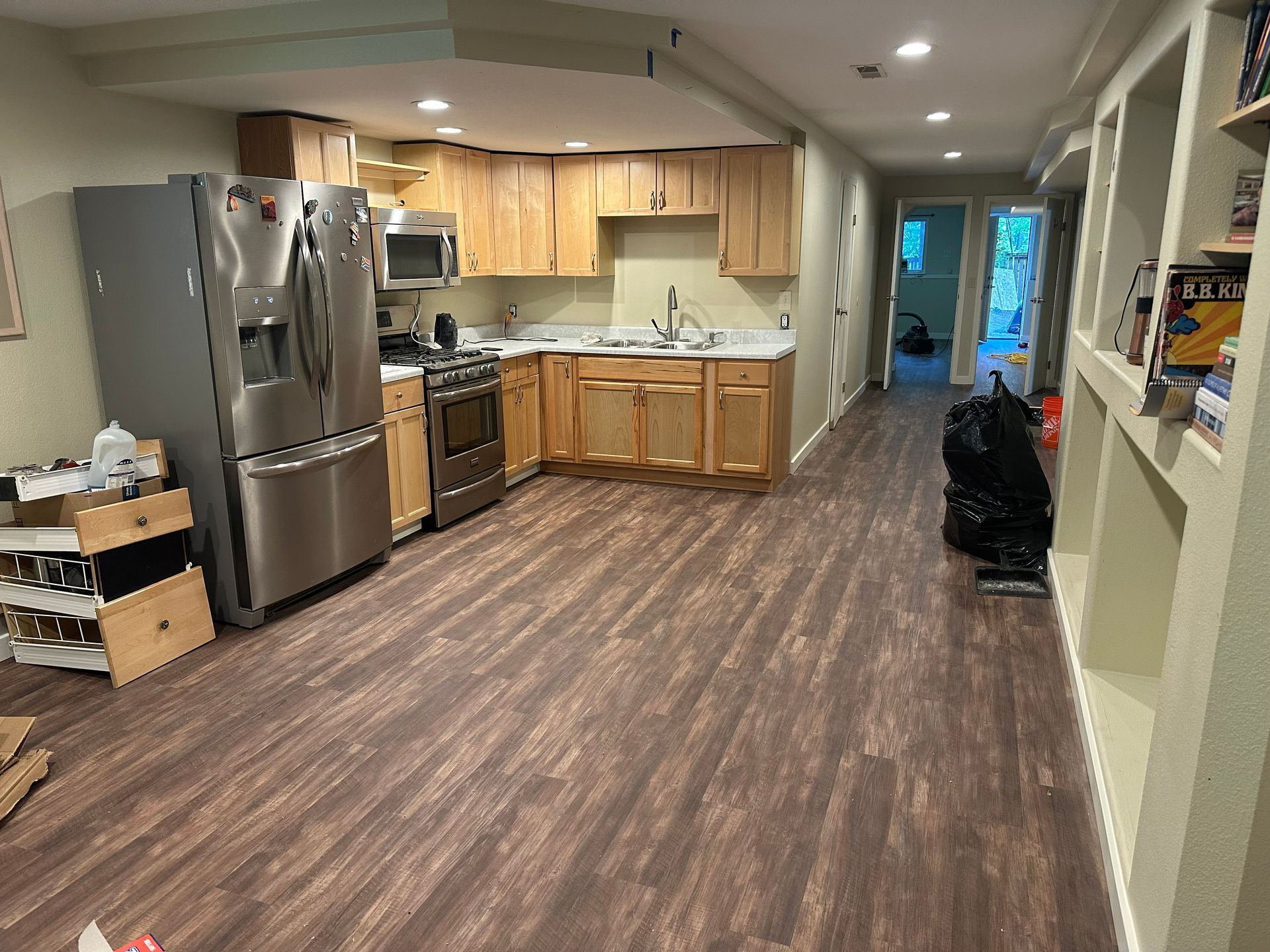 Open kitchen with stainless steel appliances, wood cabinets, and long brown floor leading to a bright doorway.