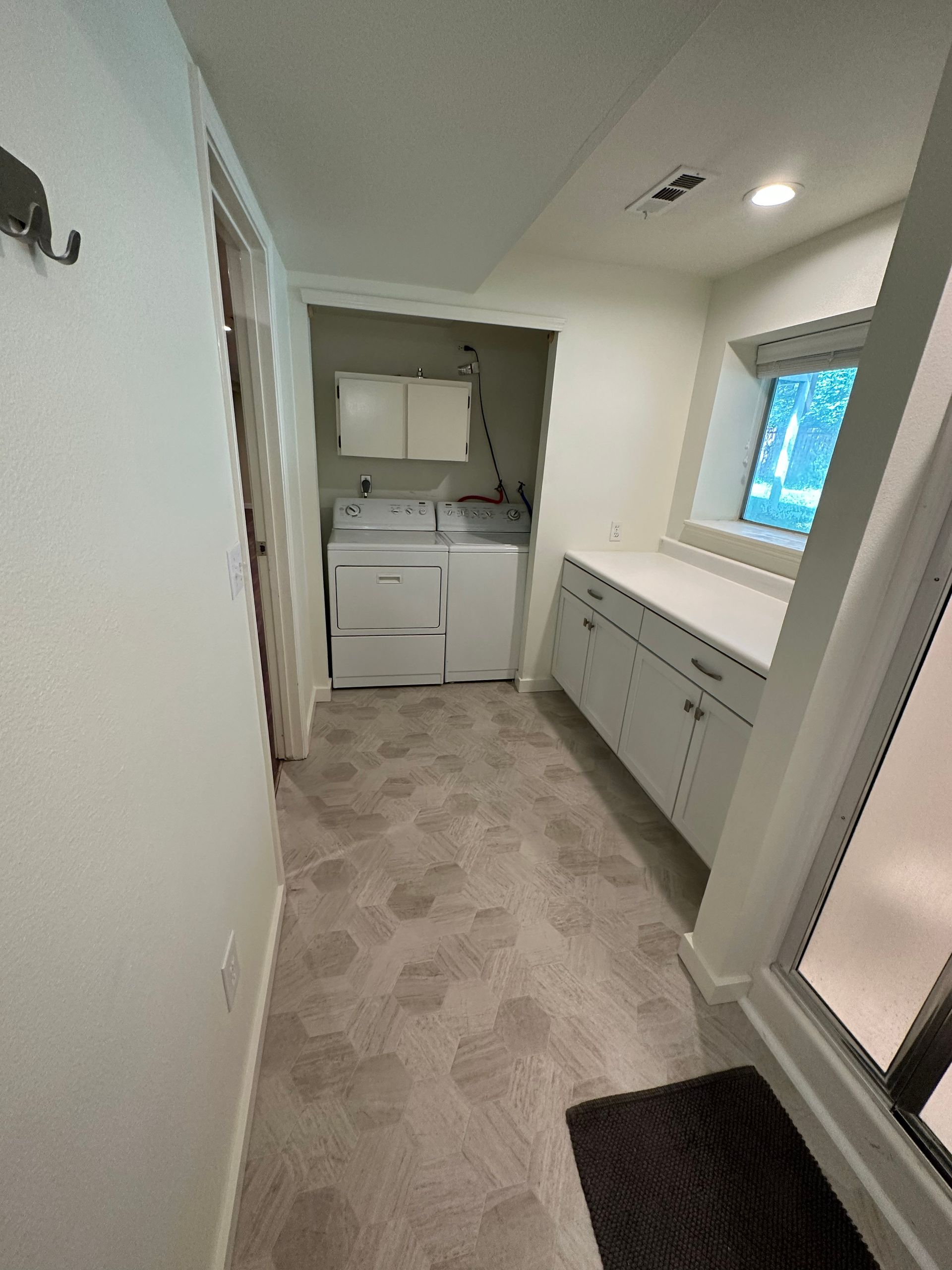 Narrow laundry room with white cabinets, countertop, window, washer-dryer, and beige carpet.