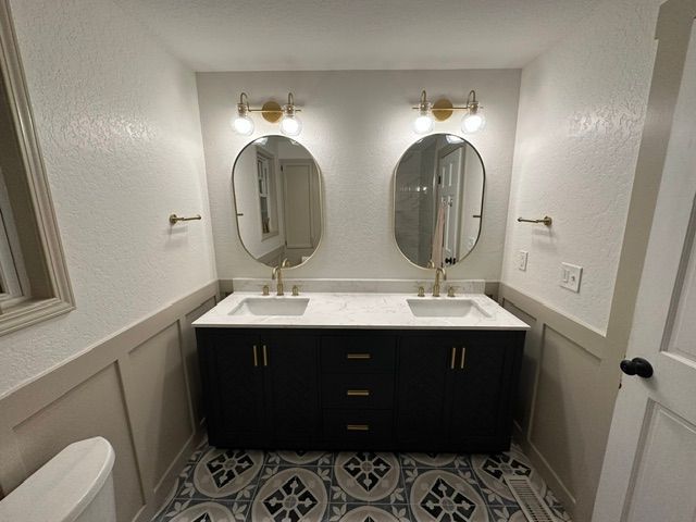Small bathroom with double vanity, oval mirrors, white walls, and patterned tile floor.