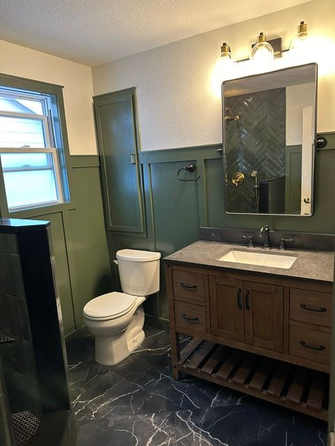 Bathroom with white toilet, dark green walls, wood vanity, mirror lights, and black marble-pattern floor.
