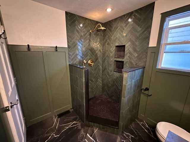 Green-tiled shower with glass door in a modern bathroom, with a window and toilet visible