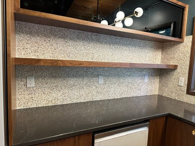 Built-in kitchen desk nook with dark countertop, speckled backsplash, shelf, and overhead mirror-light fixture