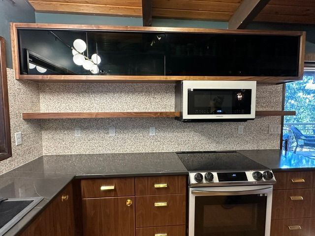 Modern kitchen with wood cabinets, granite counters, black appliance panel, shelf, and built-in microwave and stove.