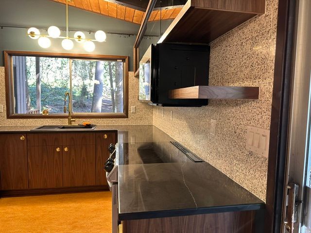 Modern kitchen interior with dark countertops, wood cabinets, patterned backsplash, and a window above the sink