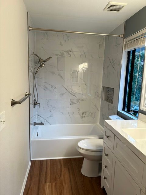 Small bathroom with bathtub, toilet, vanity, marble-look shower walls, and wood-look floor.