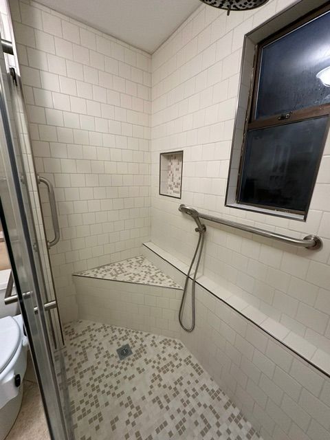Small tiled shower with grab bars, bench, and window in a bathroom.