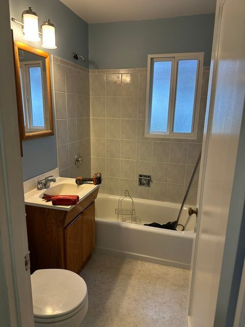 Small bathroom with a toilet, sink, bathtub, window, and tiled walls.