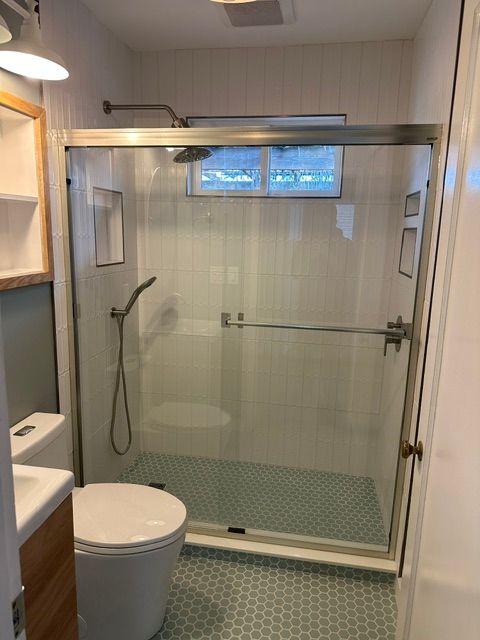 Small modern bathroom with a glass-enclosed shower, toilet, vanity edge, and window above the shower.