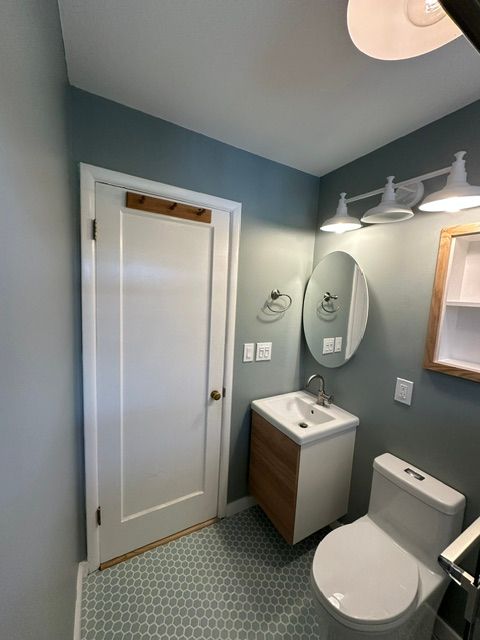 Small bathroom with white door, pedestal sink, toilet, mirror, and blue-gray walls.