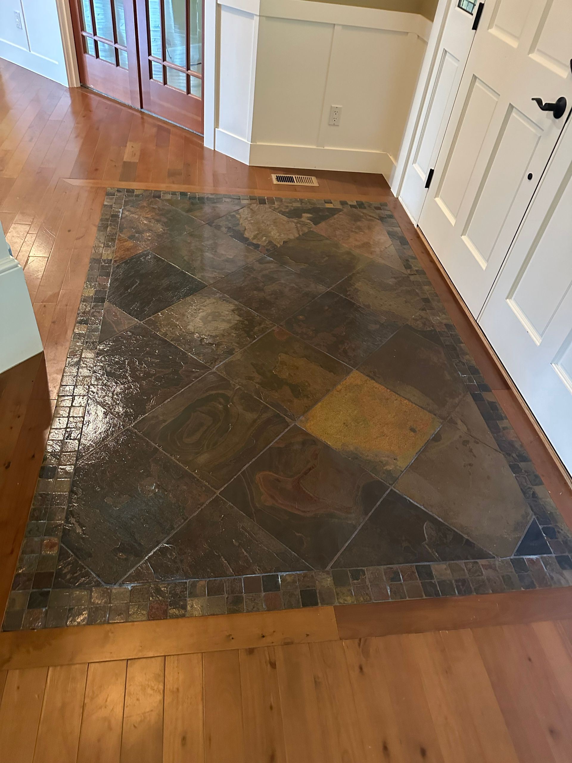 Entryway with a worn dark tile rug inset between hardwood floors and white walls, showing a tiled floor section.