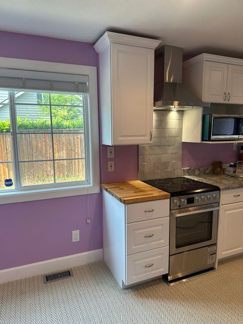 Bright kitchen with lavender walls, white cabinets, window, and stainless steel stove beside a wooden countertop.