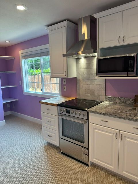 Purple kitchen with white cabinets, stainless steel stove, microwave, and a window overlooking a yard.