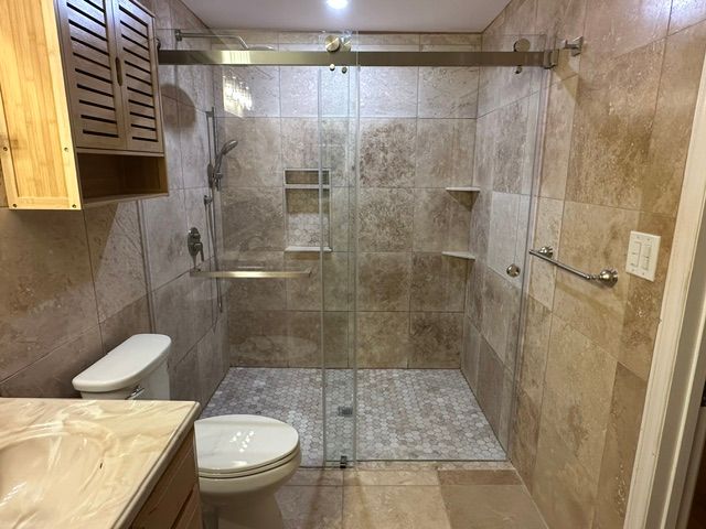 Modern beige-tiled bathroom with walk-in glass shower, toilet, and stone vanity