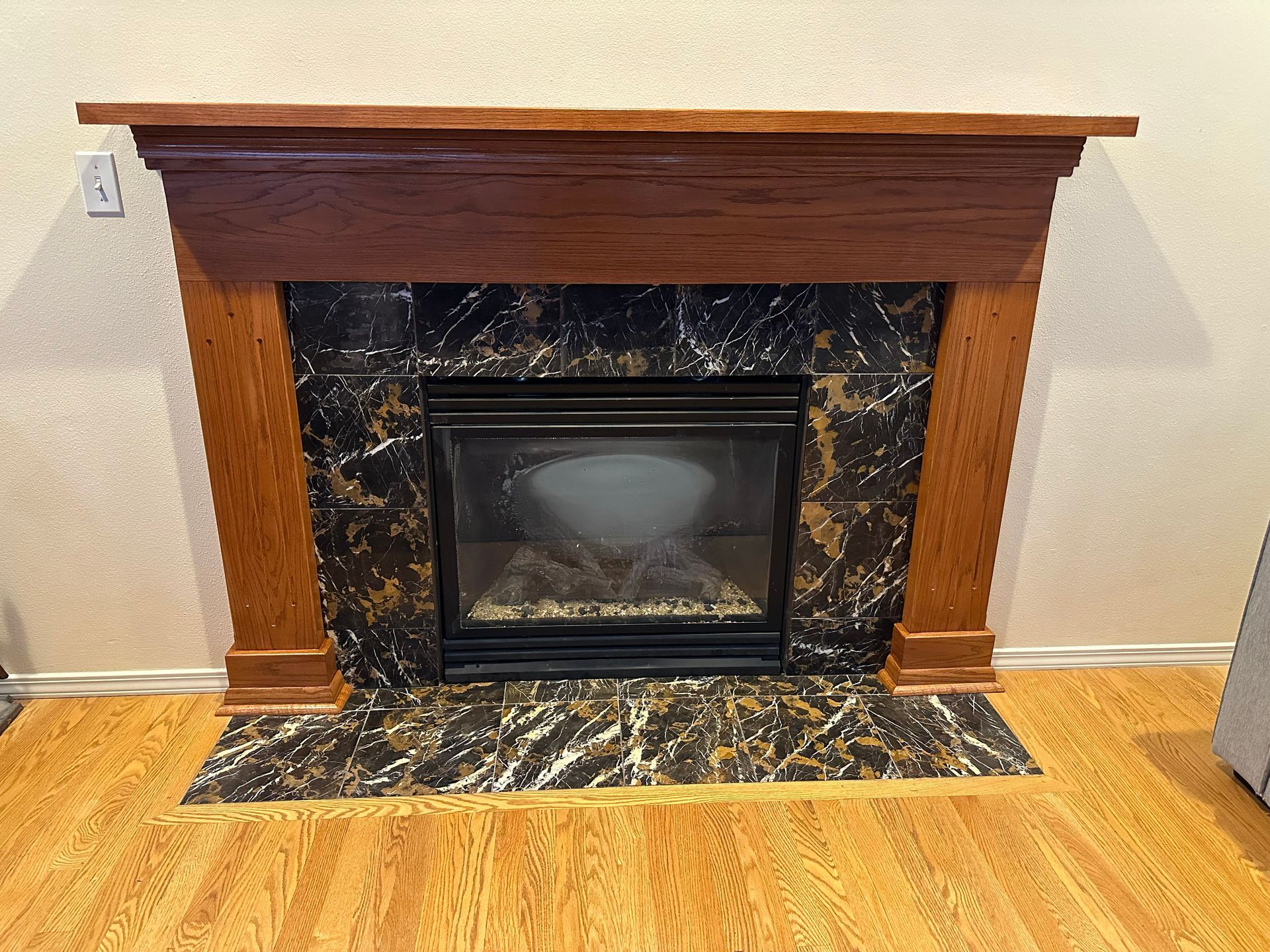 Wooden fireplace mantel with black marble surround and a glass-front insert against a beige wall.