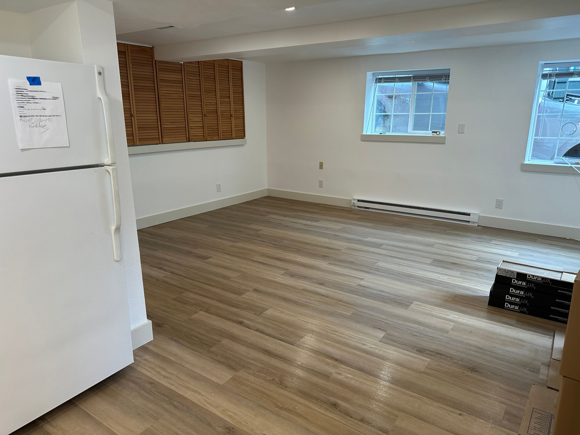 Empty room with wood floors, white walls, windows, and a white refrigerator on the left.