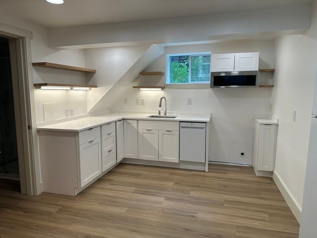 Empty modern kitchenette with white cabinets, wood flooring, and a small window above the sink