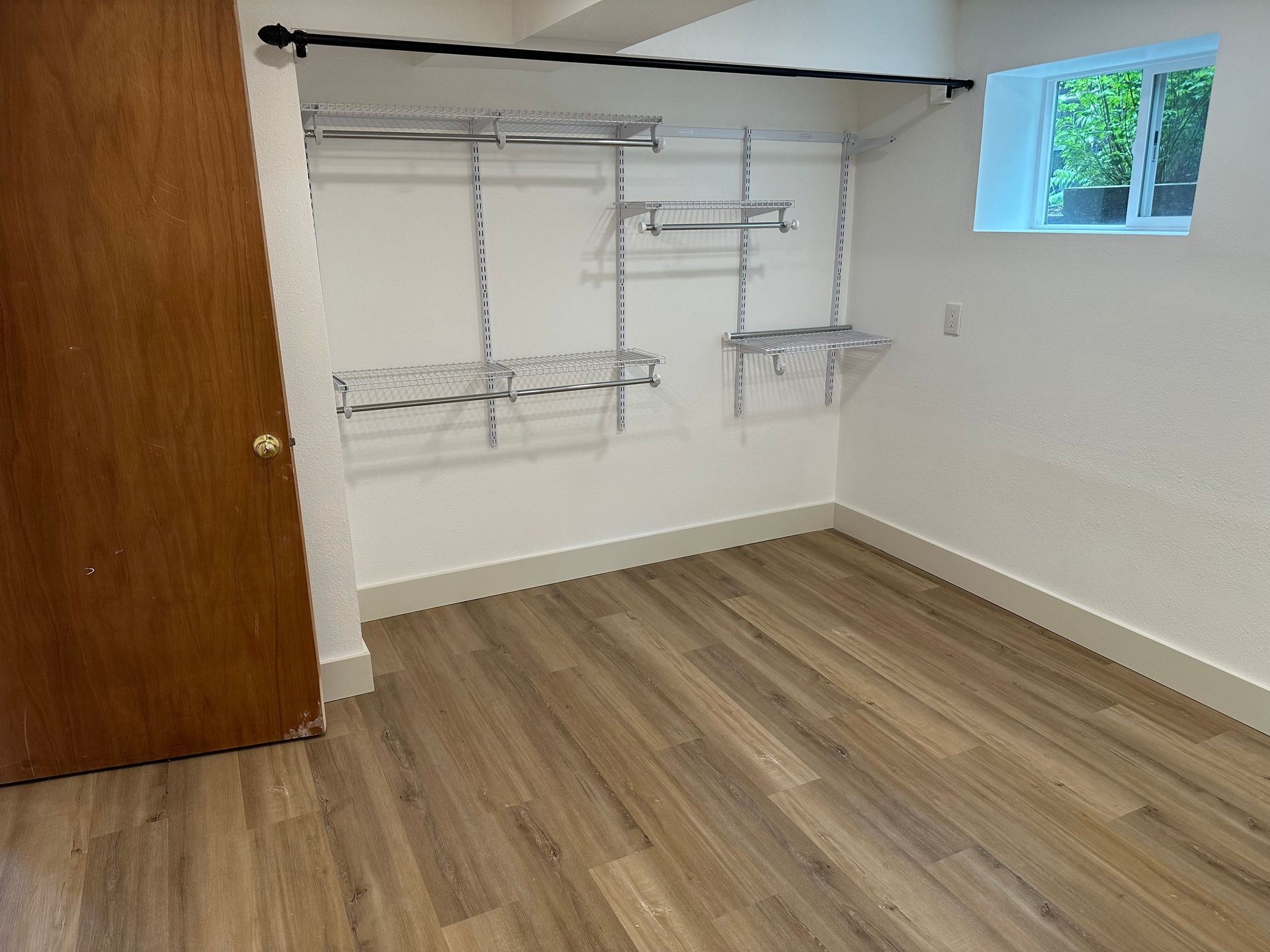 Empty closet with white walls, wood floor, hanging rods, shelves, and a small window.