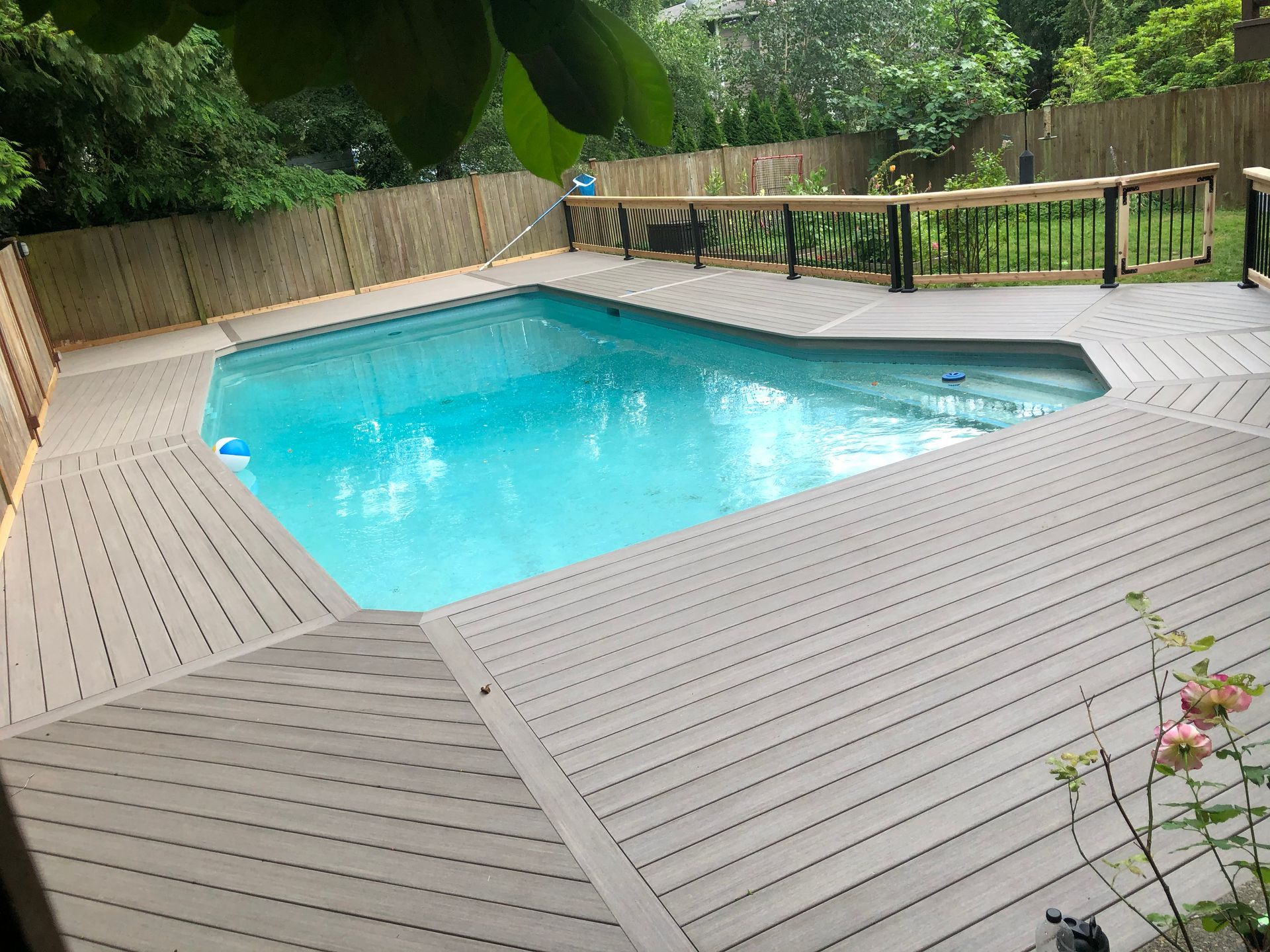 Backyard swimming pool with light blue water, wooden deck, and lush greenery around it