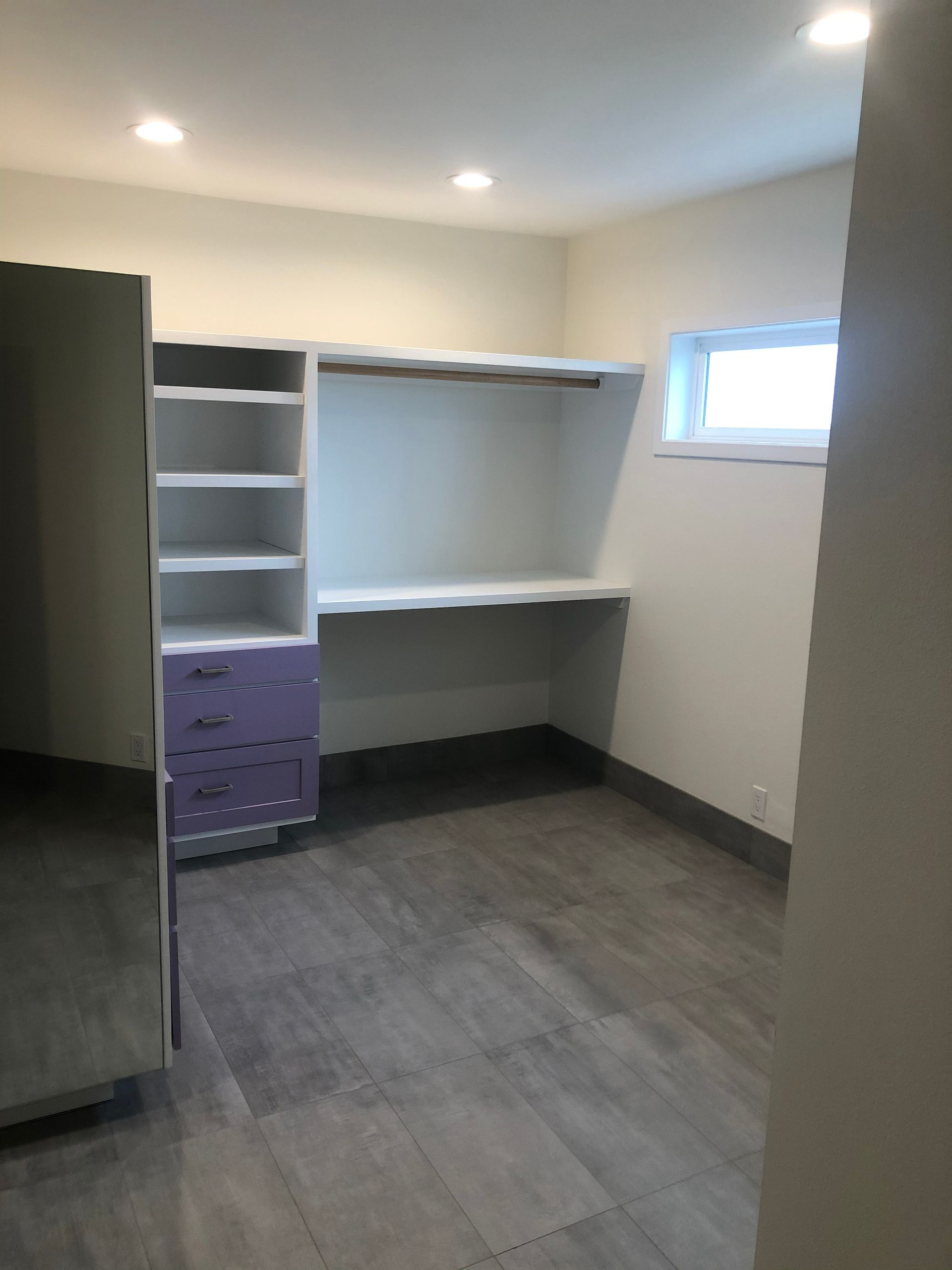 Empty walk-in closet with white shelves, purple drawers, gray carpet, and a small window.