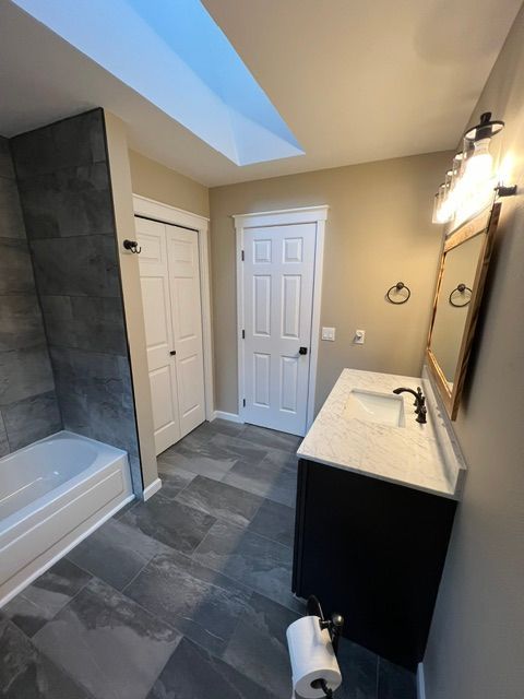 Modern bathroom with gray tile, white vanity, bathtub, toilet, and skylight.