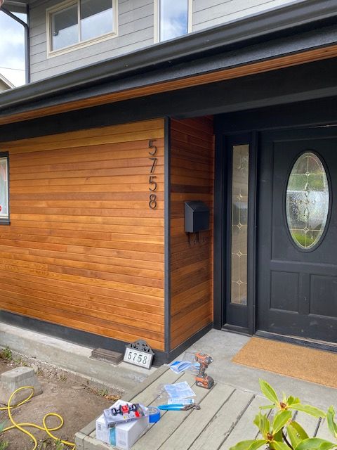 Front entrance of a house with wood siding, black door, and tools cluttering the walkway during construction.