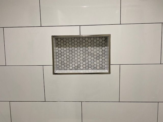White tiled wall with a small rectangular recessed opening covered by a hexagonal metal grate.