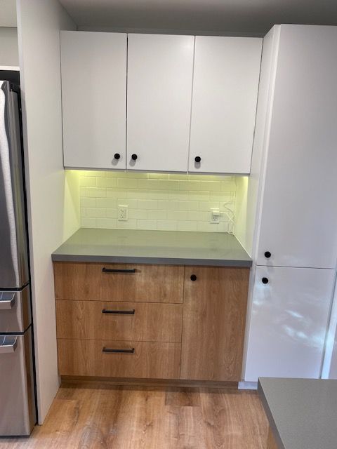 Small kitchen nook with white upper cabinets, wood lower cabinets, gray countertop, and lit pale green backsplash.