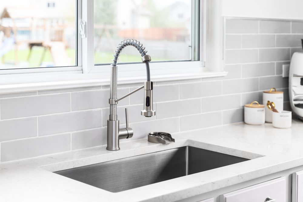 Modern kitchen sink with stainless steel faucet, white tile backsplash, and bright window light
