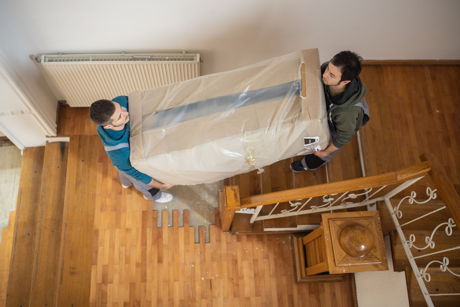 Two people carry a wrapped couch down wooden stairs, seen from above.