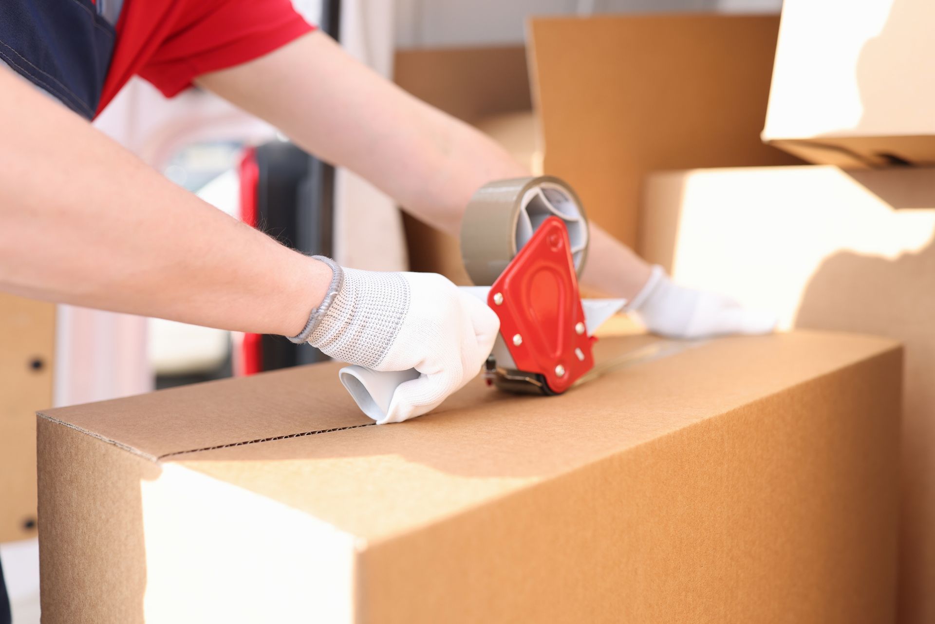 Person sealing a cardboard box with tape.  Hands wearing gloves, a tape dispenser, boxes in the background.