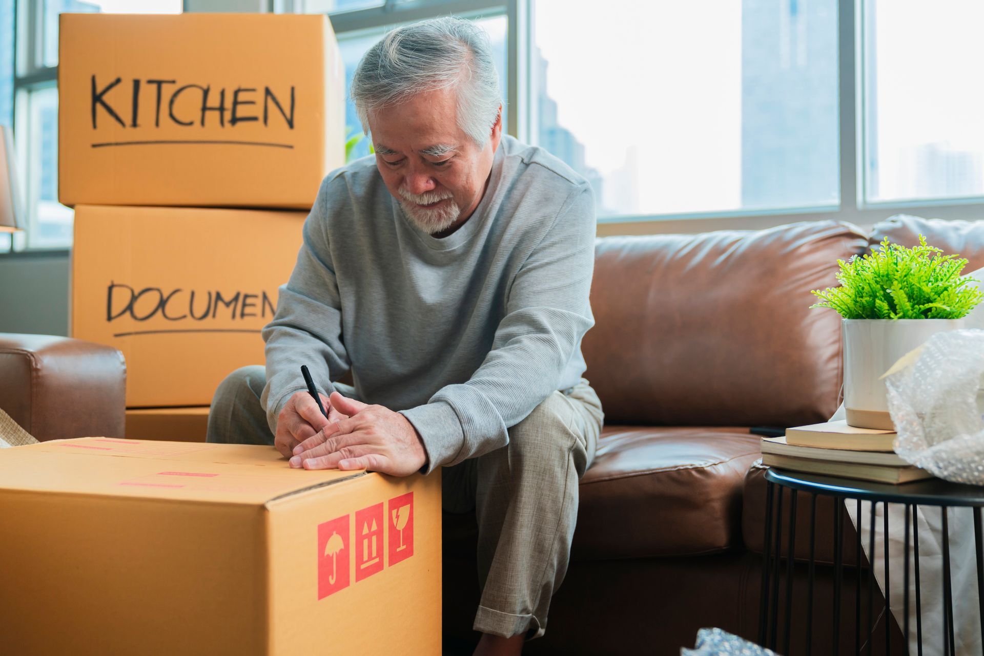 Elderly person seated on a sofa, writing on a cardboard moving box. Boxes labeled 