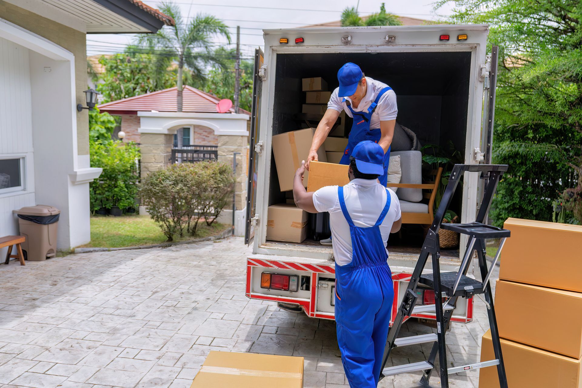 Two movers in blue coveralls load boxes into a truck, parked in a driveway.