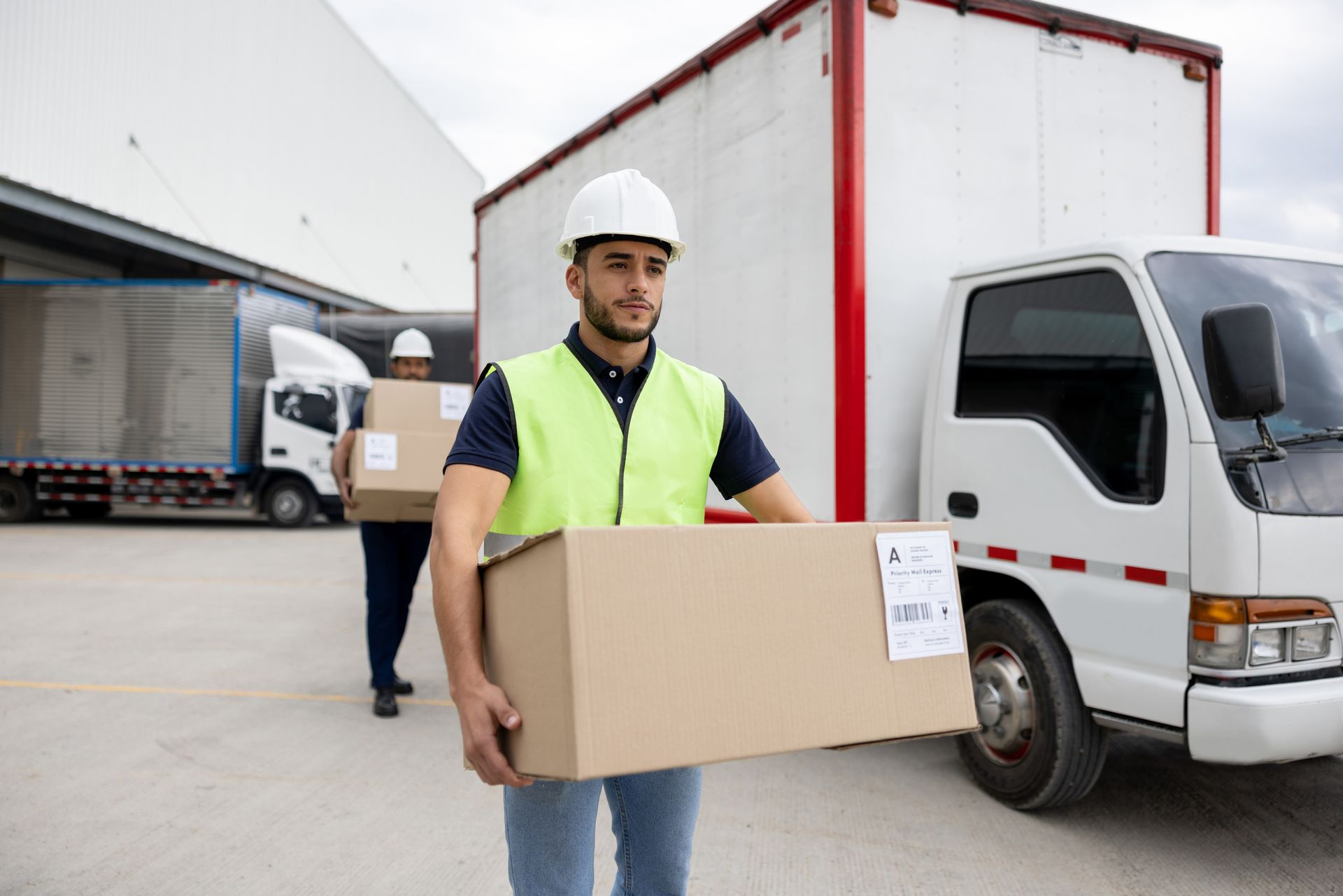 Workers carrying boxes near trucks at a loading dock; one wears a safety vest and hard hat.