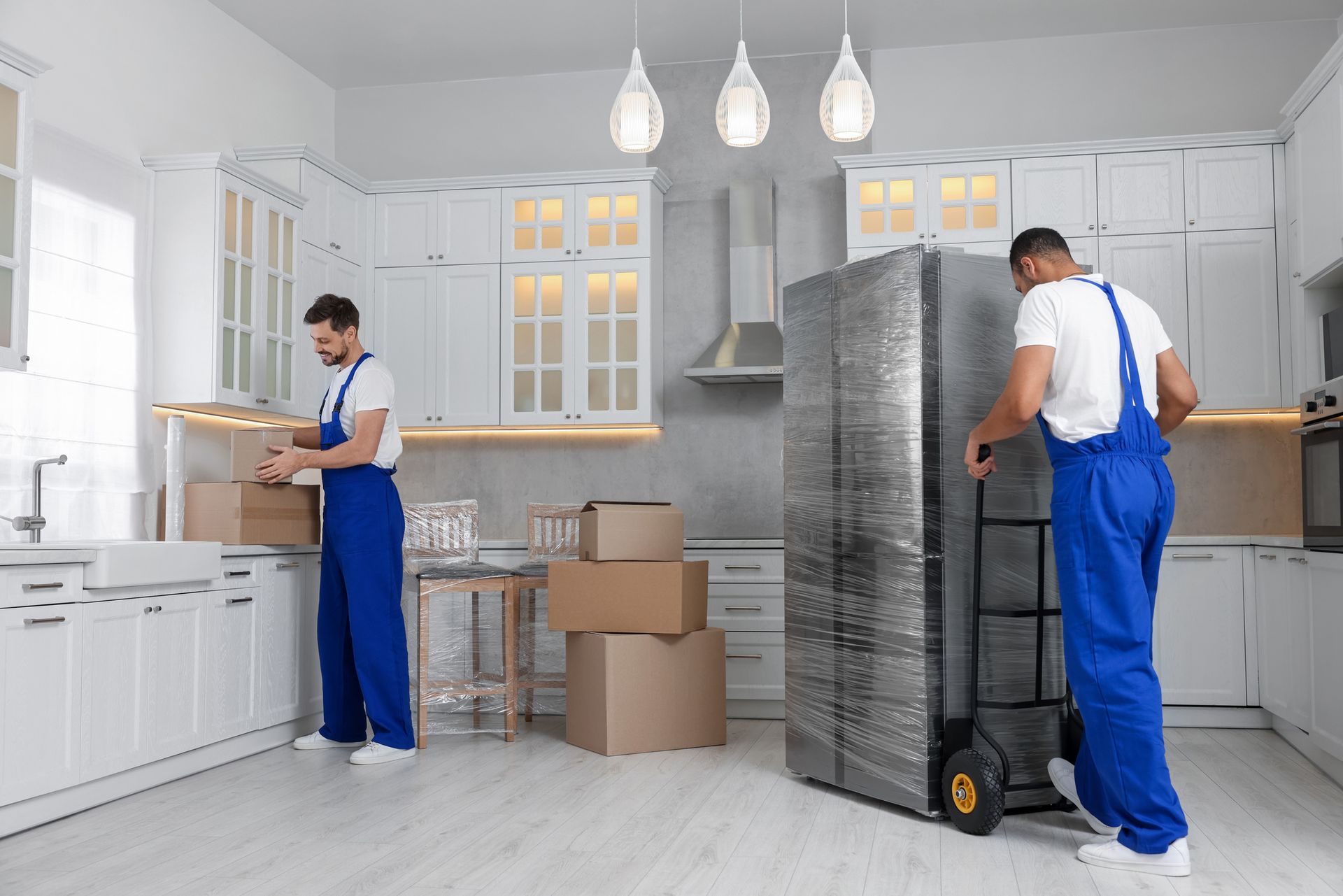 Movers in blue jumpsuits load boxes and move a wrapped refrigerator in a white kitchen.