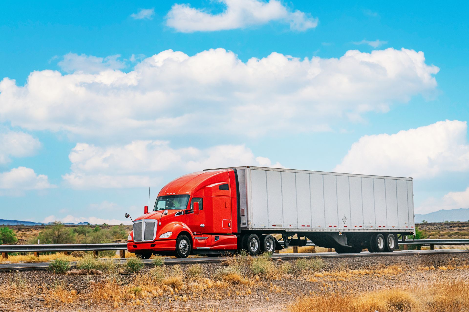 Red semi-truck driving on a highway, under a partly cloudy blue sky.