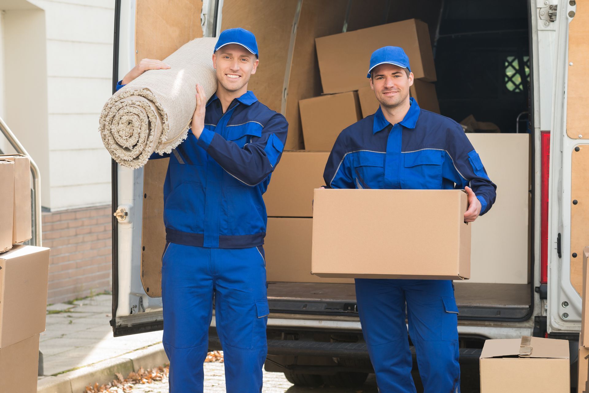 Two movers in blue uniforms carrying a rolled rug and a box in front of a moving truck.