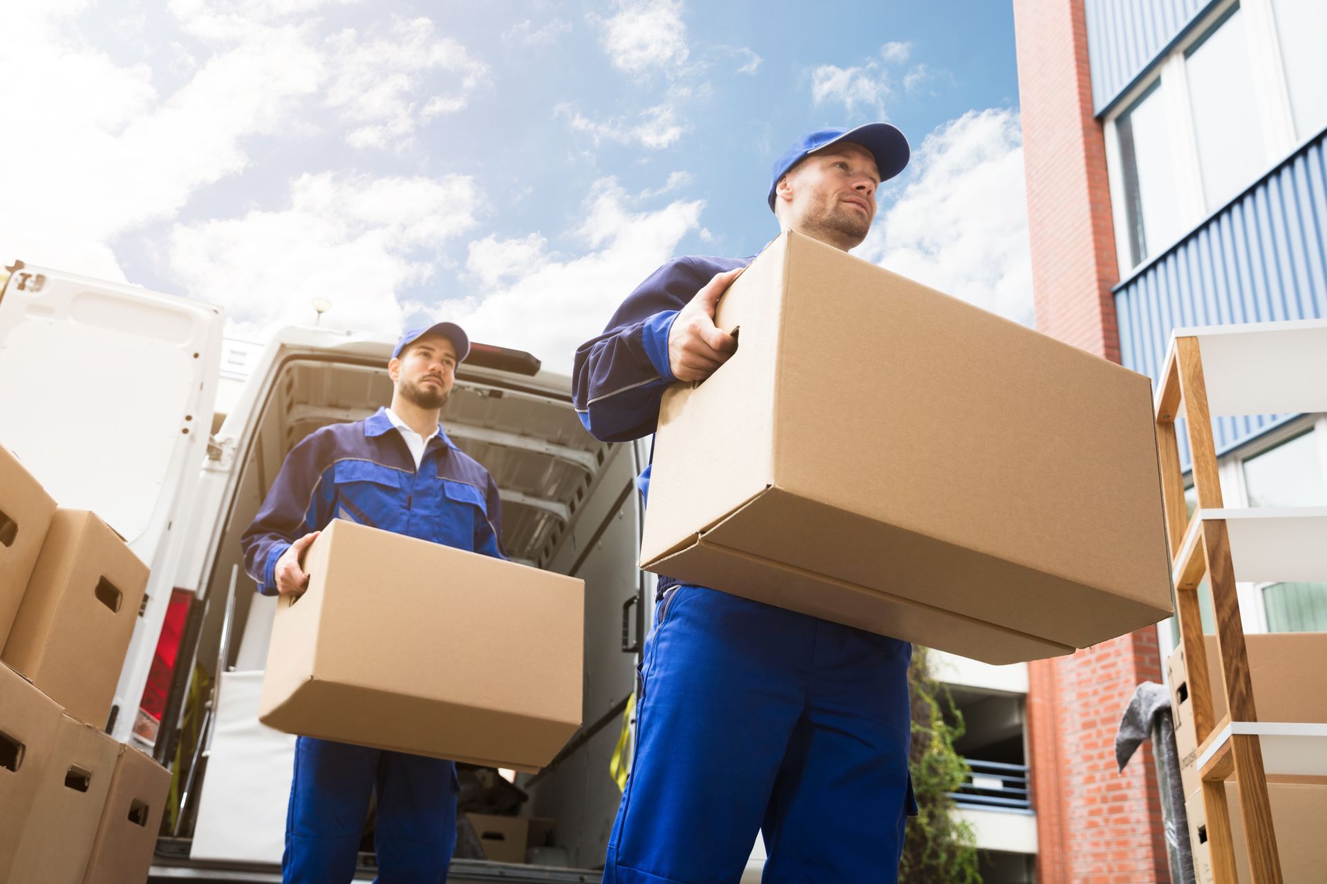 Two movers in blue uniforms carrying cardboard boxes, loading them into a truck outdoors.